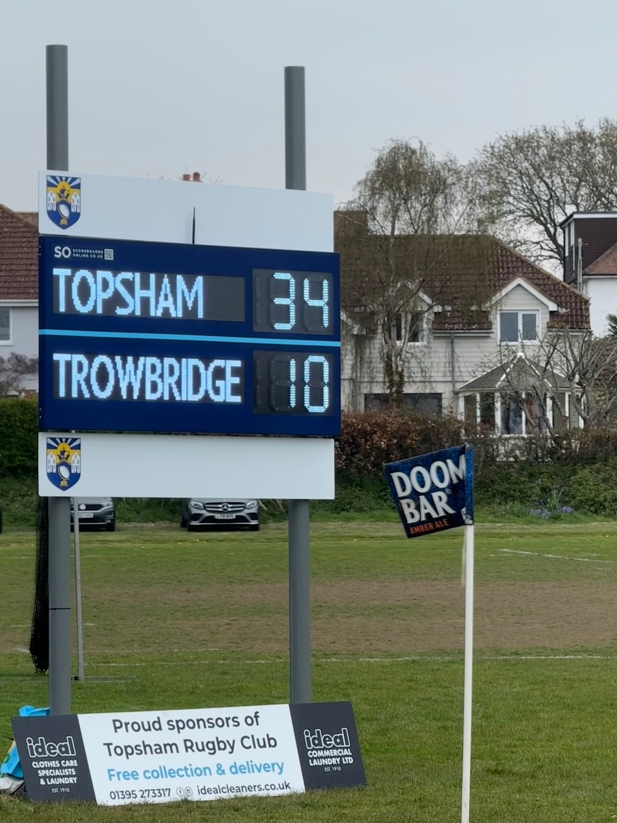 Electronic Scoreboard with editable LED names, displaying Topsham vs Trowbridge with a rugby club sponsorship sign in the background.